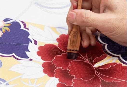 筆で赤い牡丹柄の生地に色を入れる職人の手元の写真